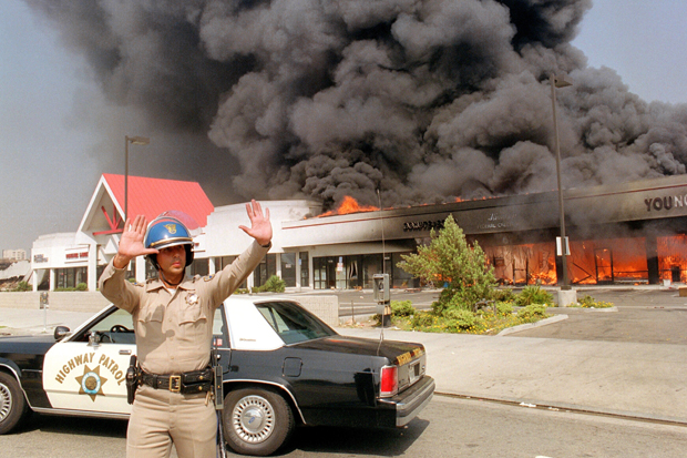 A California Highway patrolman directs raffic around a shopping center engulfed in flames in Los Angeles, 30 April 1992. Riots broke out in Los Angeles, 29 April 1992, after a jury acquitted four police officers accused of beating a black youth, Rodney King, in 1991, hours after the verdict was announced. (Photo credit should read CARLOS SCHIEBECK/AFP/Getty Images)